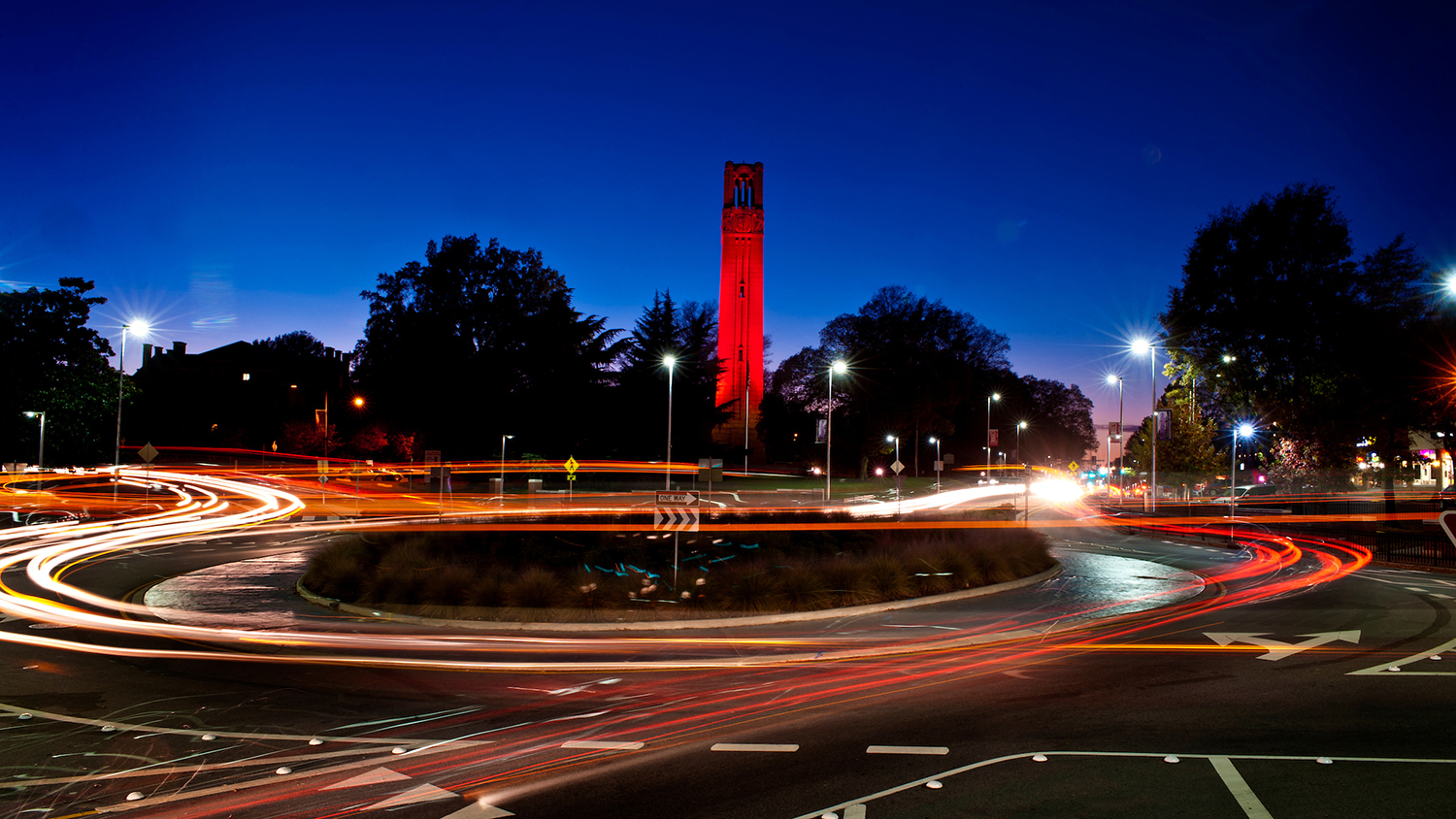 NC State belltower in red light with timelapse traffic headlights in foreground.