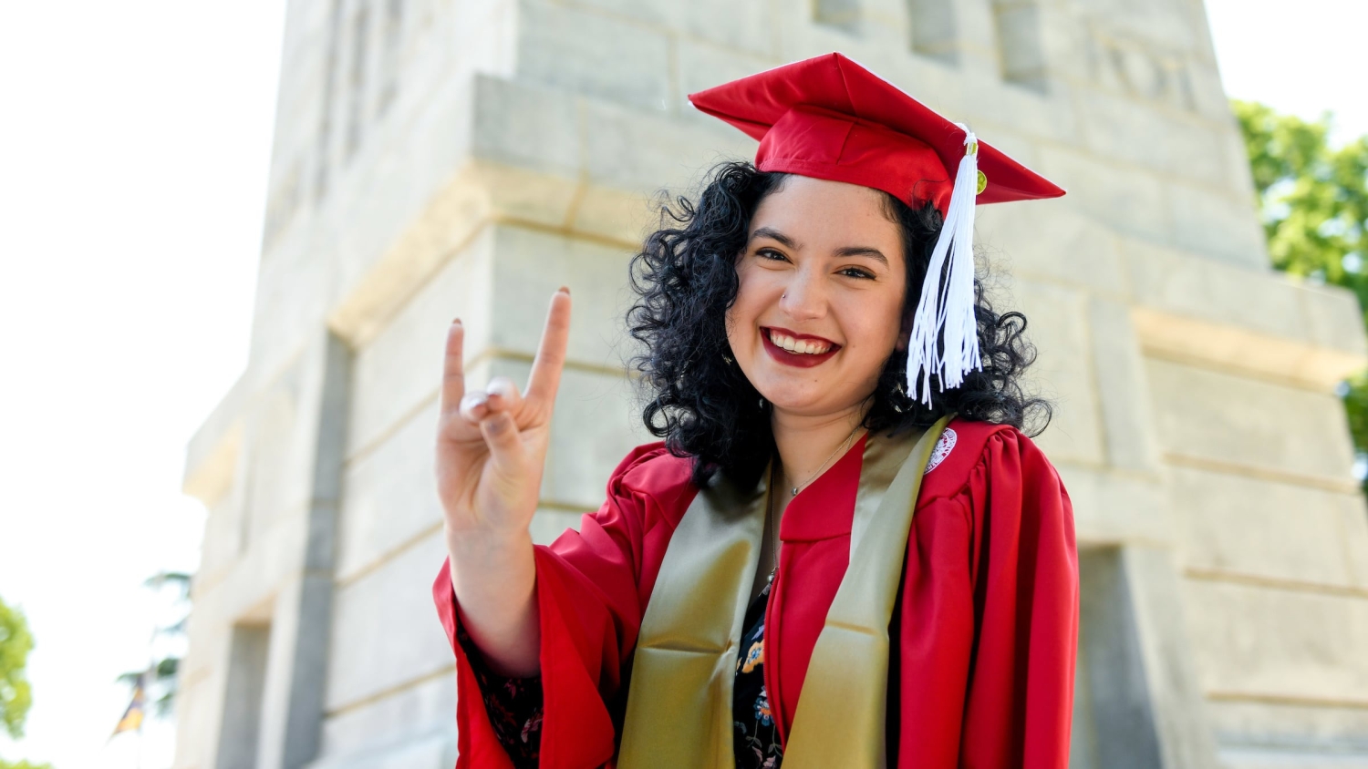 A student in a red cap and gown makes the Wolfpack sign in front of the NC State Belltower.